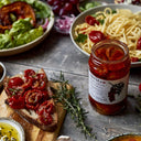 A jar of Odyssey Sunflower Tomatoes on the cobb on a rustic wooden table, surrounded by prepared food including pasta, salad, and toast with sunflower tomatoes.