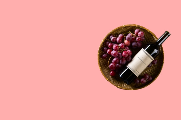 A dark glass bottle with a blank cream label rests on a bed of red grapes inside a round woven basket, set against a solid pink background.