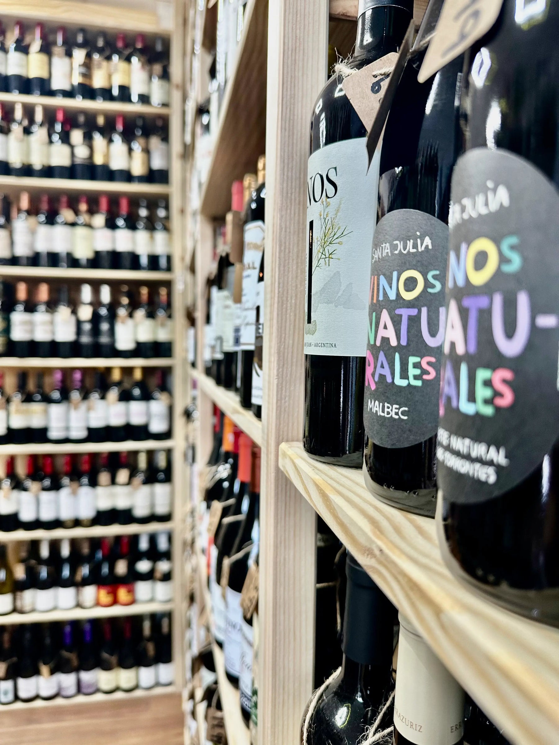 A close-up of dark wine bottles on a wooden shelf in a wine store, with shelves of other bottles blurred in the background. The front bottle has a black label with colorful text reading 'NOS NATURALES' and another visible label reads 'VIR JULIA'.