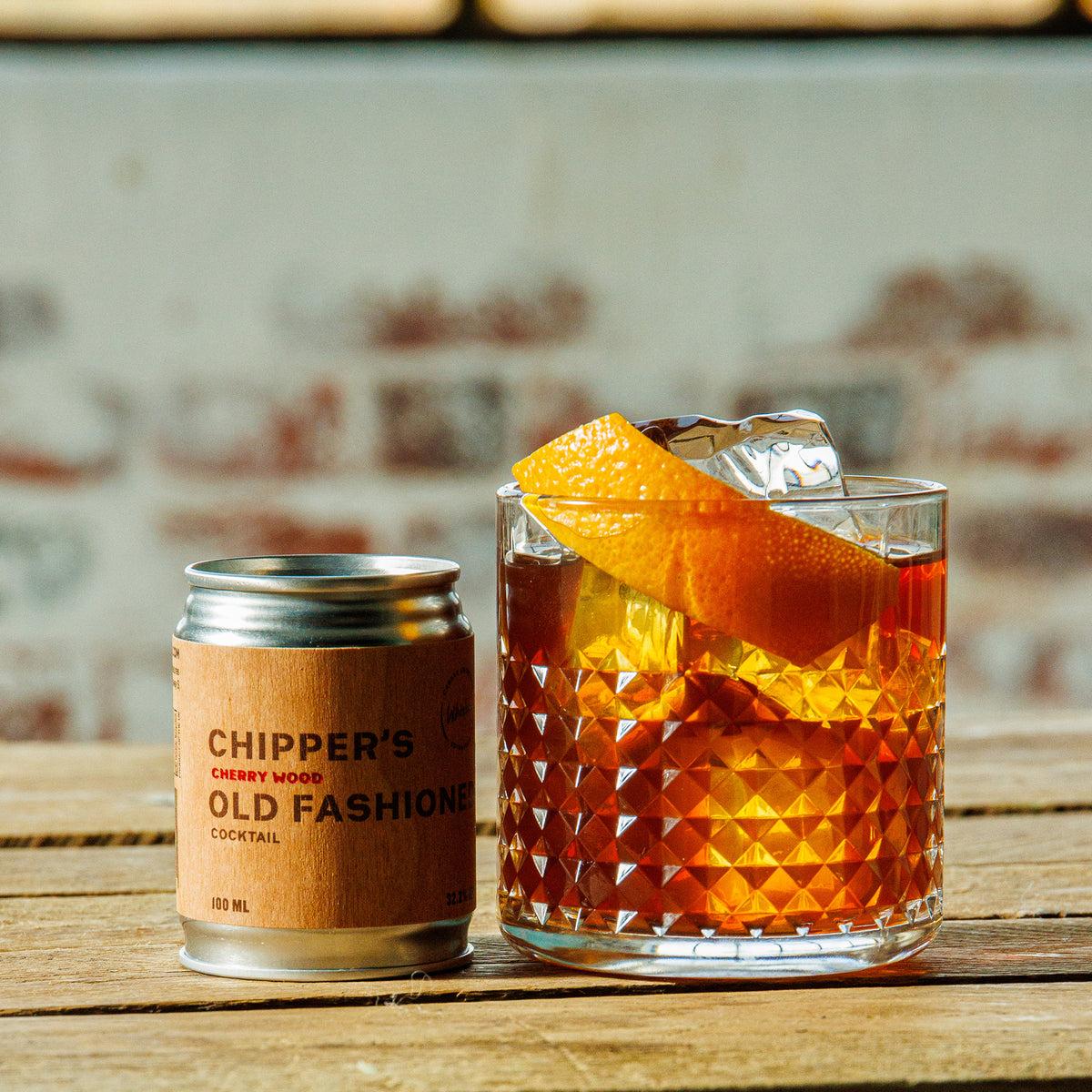 Cocktail in a glass with an orange slice and ice cube, next to a can of 'Chipper's Cherry Wood Old Fashioned Cocktail' on a wooden surface.
