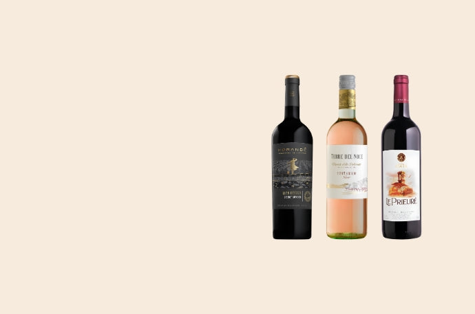Three wine bottles arranged side by side on a plain warm beige background: a dark red wine bottle, a pale pink wine bottle, and a dark red wine bottle with a white label.